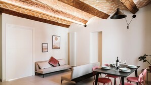 A modern living room with a sofa, dining table, and chairs. There is a radiator, a framed picture on the wall, and a plant in the corner. - Vicolo al Castello - Langhe e Monferrato Suites (Costigliole d'Asti)