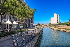 A riverside walkway with a wooden deck, metal railing, and a building with a modern design in the background. - Spacious 2Bed Harbor Haven | V&A Waterfront Canals (Cape Town)