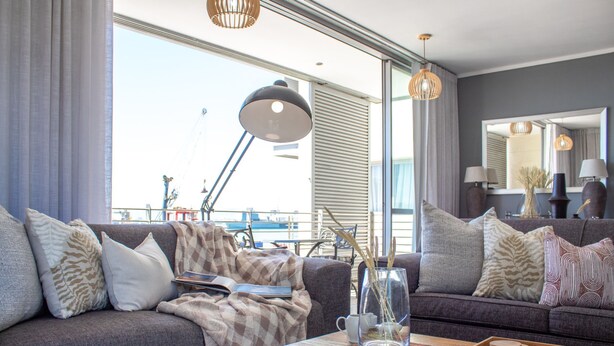 A modern living room with a grey sofa, a coffee table with a board game, and a large window with curtains. - Spacious 2Bed Harbor Haven | V&A Waterfront Canals (Cape Town)