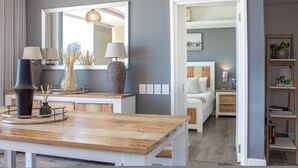 A modern living room with a wooden coffee table, two lamps, and a large mirror reflecting the room. A doorway leads to a bedroom with a bed and a nightstand. - Spacious 2Bed Harbor Haven | V&A Waterfront Canals (Cape Town)