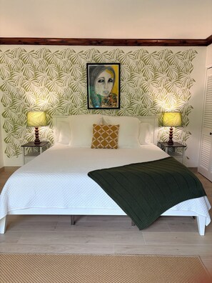 Une chambre à coucher avec un lit, deux lampes de chevet, un papier peint à motifs, une œuvre d’art encadrée au-dessus du lit et un plaid vert.