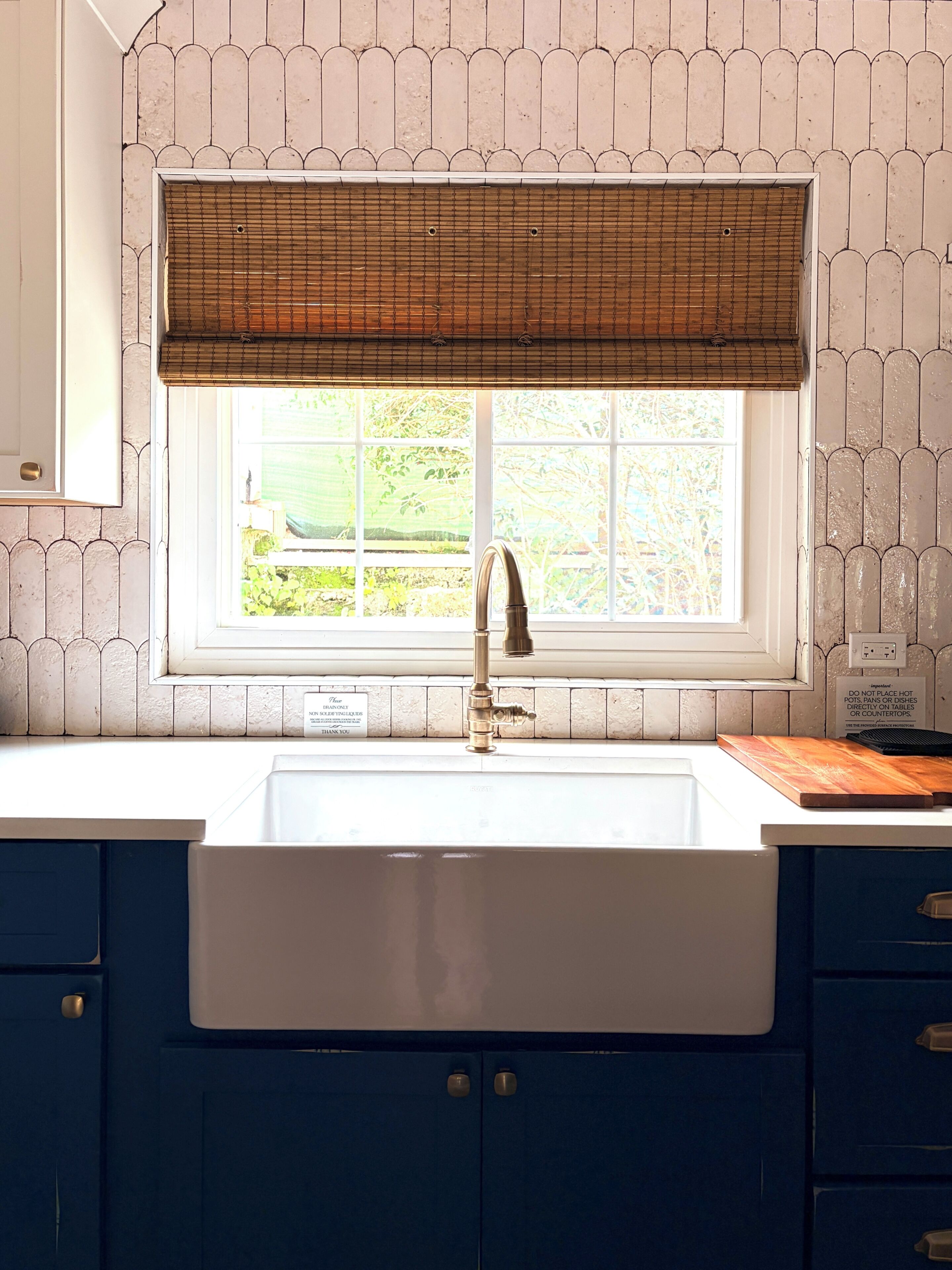 A kitchen with a white sink, a window with a bamboo blind, and blue cabinets.