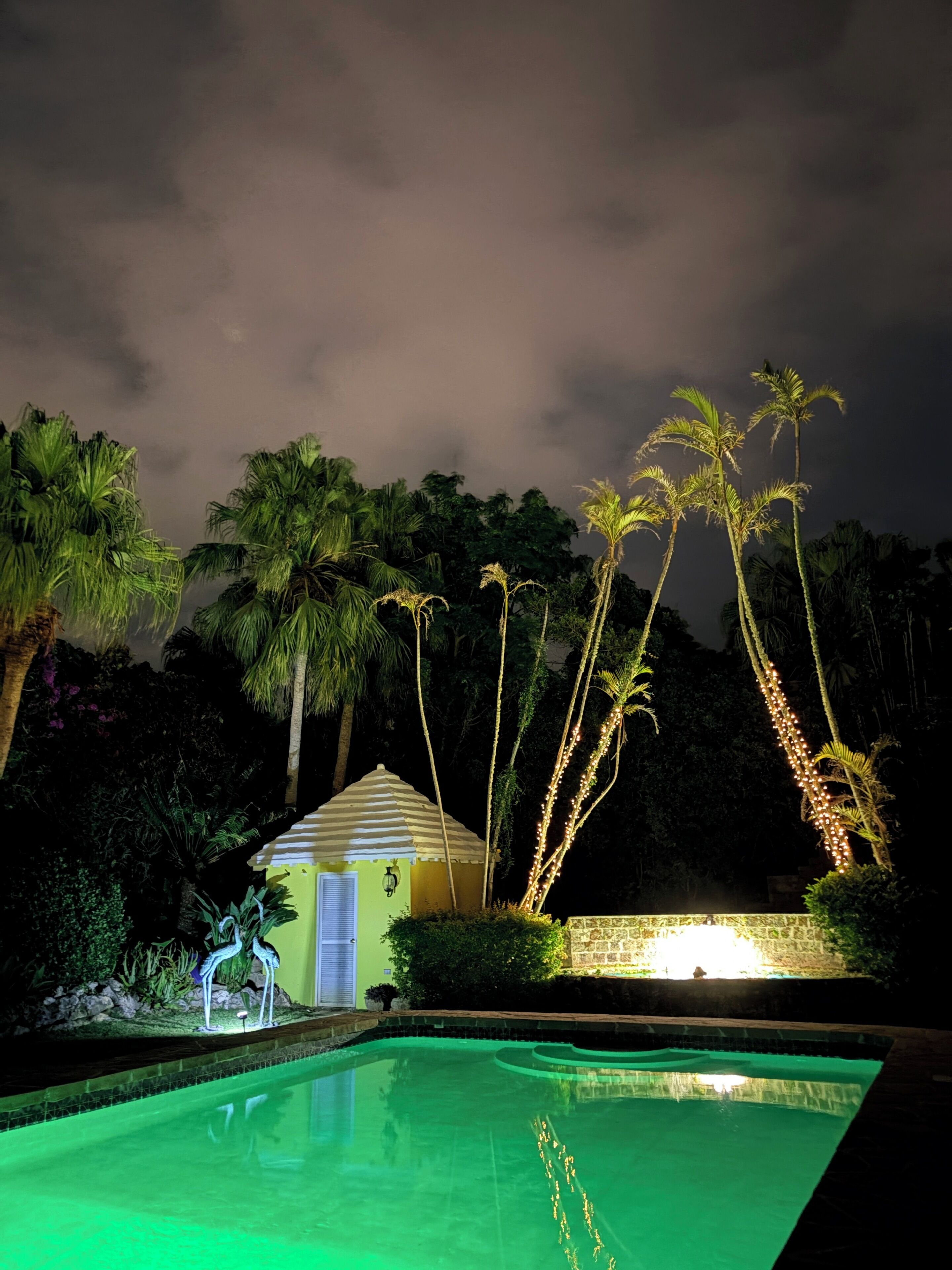 A nighttime view of a swimming pool area with a small gazebo and palm trees.