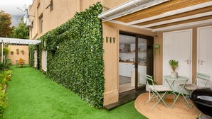 Un patio con césped artificial, una pared verde, una zona de comedor con sillas y una mesa, y una parrilla de barbacoa.