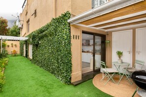 Ein Wohnhaus mit Rasen, einer mit Efeu bewachsenen Mauer, einer Schiebetür zu einer Küche und einer kleinen Terrasse mit Tisch und Stühlen.
