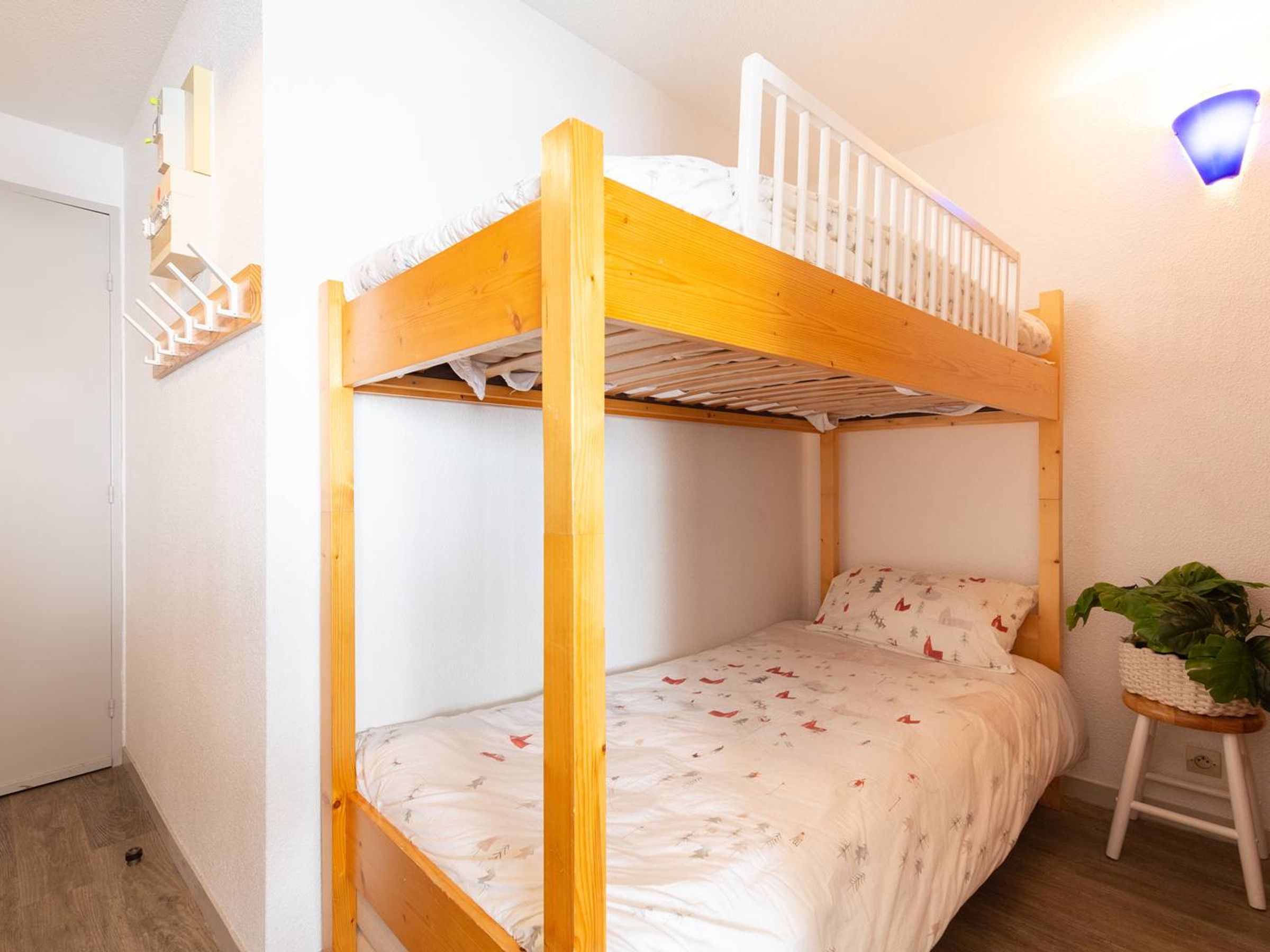 A bunk bed with a wooden frame, a lower bed with a patterned bedspread, and a small stool with a plant.