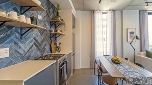 A modern kitchen with a herringbone tile backsplash, wooden shelves, and a dining area with a long table. - Two Bed | Sea Point | Rooftop Pool (Cape Town)
