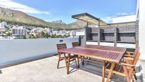 A rooftop terrace with a wooden table and chairs, a pergola, and a view of a cityscape with mountains in the background. - Two Bed | Sea Point | Rooftop Pool (Cape Town)