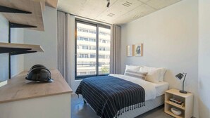 A modern bedroom with a large bed, a desk with a helmet, a nightstand with a lamp, and a window with a view of a building. - Two Bed | Sea Point | Rooftop Pool (Cape Town)