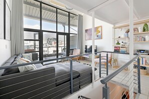 A modern living room with a large window, grey sofa, and a staircase leading to an upper level. - Two Bed | Table Mountain Views | Central (Cape Town)