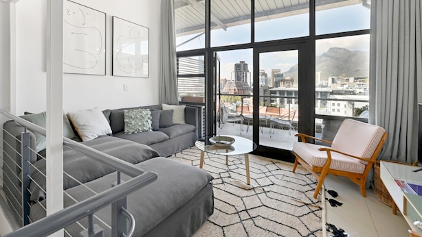 A modern living room with a grey sofa, a small round table, and a wicker chair. A large window offers a city view. - Two Bed | Table Mountain Views | Central (Cape Town)