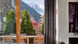 A modern room with a large window overlooking a mountain and a red bus. There is a wooden table and chairs, a black chair with a heart design, and a white cup on the table. - Chalet Cristal (Chamonix)