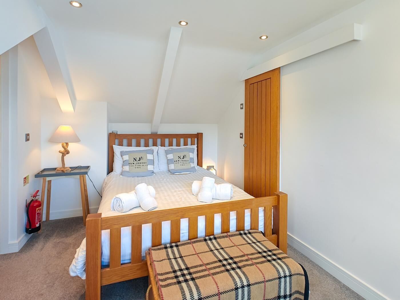 A bedroom with a wooden bed, two bedside tables, a bench, a fire extinguisher, and a door.