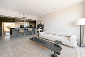 Ein modernes Wohnzimmer mit Sofa, Couchtisch und Barhocker. Im Hintergrund ist eine Küche erkennbar.