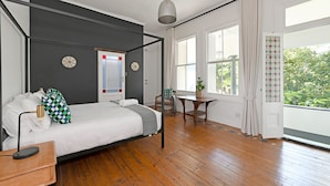 Un dormitorio con cama con dosel, suelo de madera, una mesita con una planta y una ventana con cortinas.