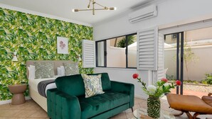 A room with a green sofa, a bed with white and patterned pillows, a glass coffee table, and a tropical wallpaper. - Stylish Sea Point Studio in Secure Complex + Patio (Cape Town)