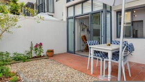 A modern outdoor patio with a white table and blue and white patterned chairs, surrounded by a gravel area and potted plants. - Stylish Sea Point Studio in Secure Complex + Patio (Cape Town)
