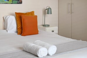 Ein ordentlich bezogenes Bett mit weißen und orangefarbenen Kissen, einer grauen Lampe und einem weißen Nachttisch.
