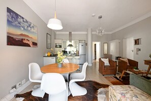 A modern living room with a dining area, kitchen in the background, and a large painting on the wall. - Charming Sea Point 1 Bed with Leafy Patio (Cape Town)