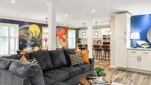 A modern living room with a grey sectional sofa, a dining area with a large mural, and a kitchen with white cabinets and a blue accent wall. - Pool Table + Hot Tub - Raleigh Retreat (Raleigh)