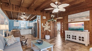 A spacious living area with a wooden ceiling, a ceiling fan, a television, and a dining area with a kitchen in the background.