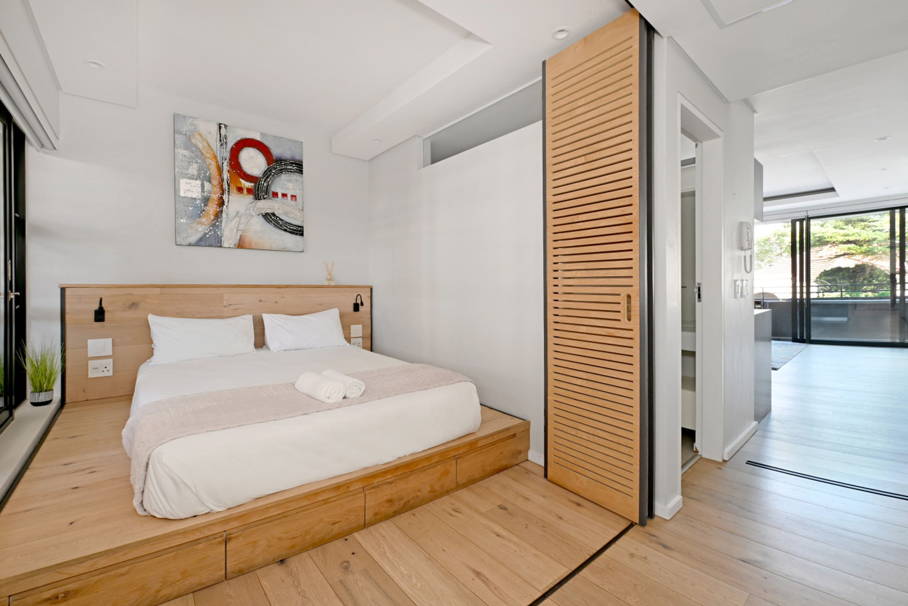 Een moderne slaapkamer met een bed op een houten platform, een groot abstract schilderij en een houten schuifdeur naar een balkon.