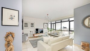 Un soggiorno moderno con un divano bianco, un tavolino da caffè in vetro e una TV sullo sfondo. Si notano quadri appesi alla parete, un'ampia finestra con vista e una scultura in legno decorativa.