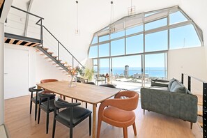 A spacious living area with a large window overlooking the sea, a wooden dining table with chairs, and a modern sofa. - Spacious 3 Bedroom Villa | BBQ | Pool | Ocean View (Cape Town)