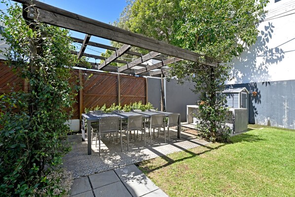 Een terras met een tafel en stoelen, een houten pergola en een kleine tuin met een hek en begroeiing.