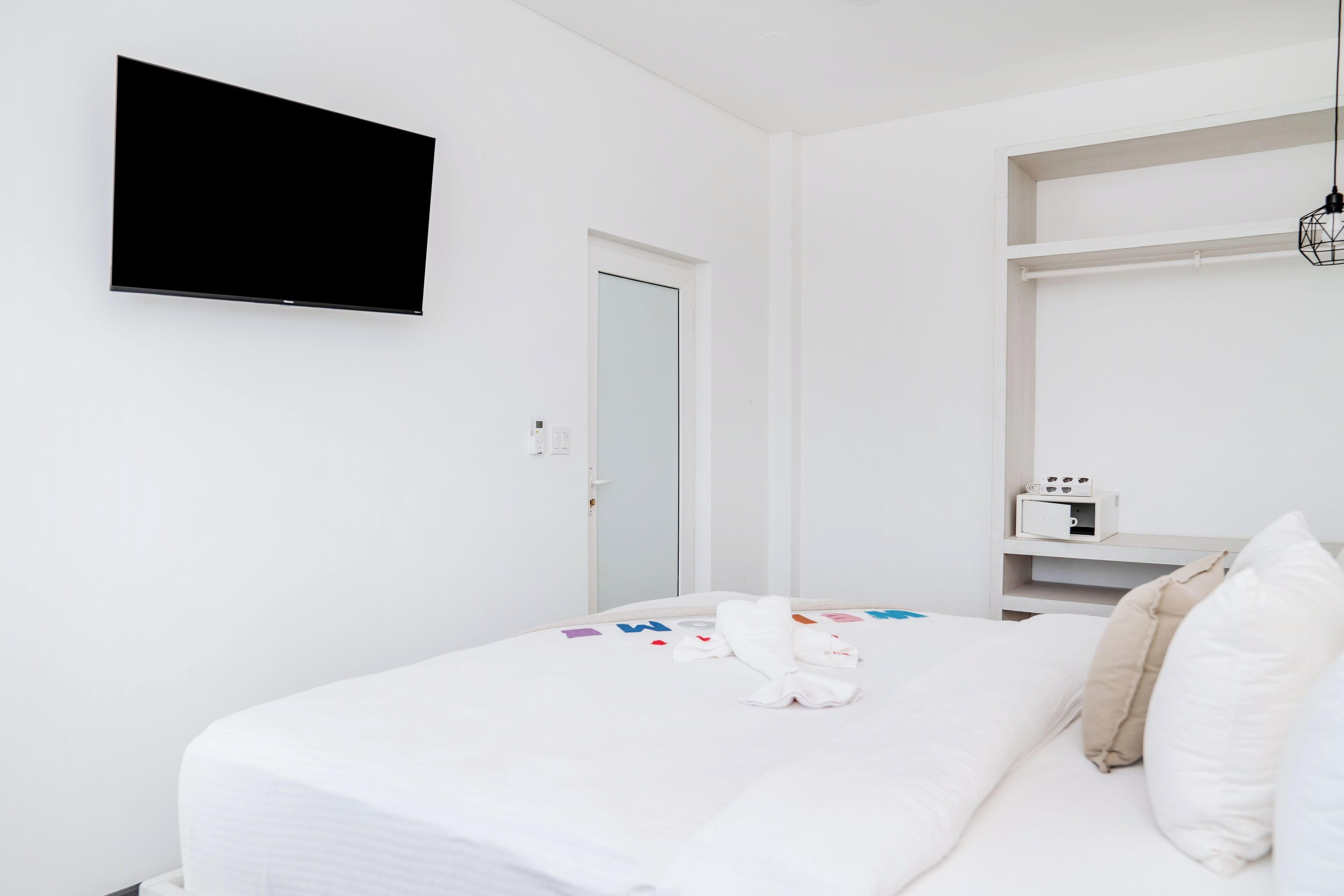 A modern hotel room with a large bed, a flat-screen TV mounted on the wall, a bedside table with a lamp, and a wardrobe.