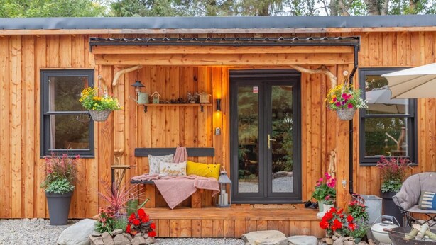 A wooden cabin with a front porch, a bed with pillows, a small table, and hanging flower baskets. - 2 Bed in Invergordon (oc-c33073) (Invergordon)