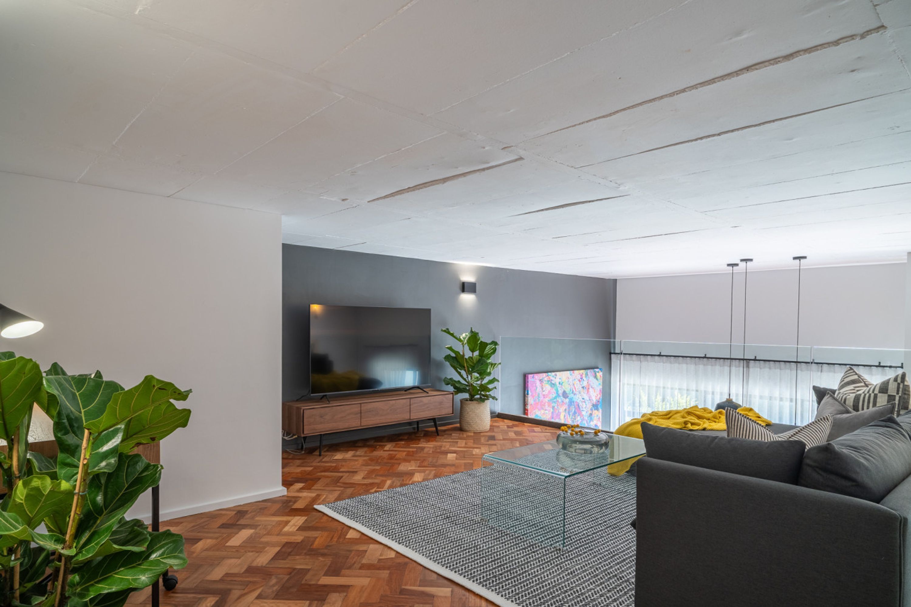 A modern living room with a herringbone floor, a glass coffee table, a grey sofa, a TV, and a large plant.