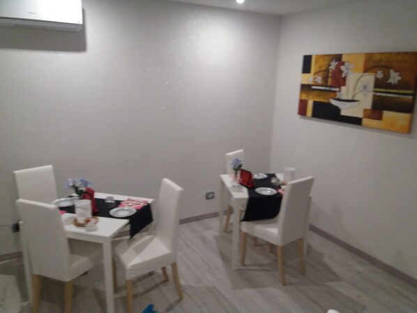 A dining area with two white tables, each set with plates, glasses, and a red napkin. The room has white walls and a painting on the wall.