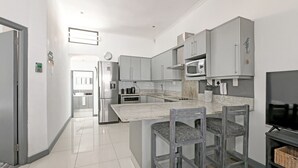 A modern kitchen with grey cabinets, a stainless steel refrigerator, a microwave, and a TV. There are two wooden chairs with cushions. - Secure Sea Point | Spacious | Three Bedroom house (Cape Town)