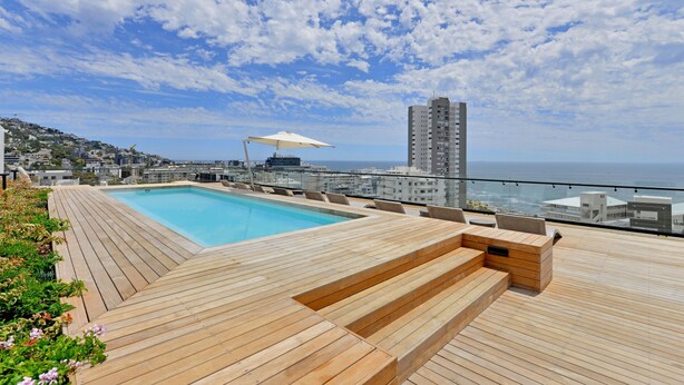 A rooftop pool area with wooden decking, a lounge with seating, and a view of the ocean and cityscape. - Station house | Studio Apartment | Gym |Rooftop Pool (Cape Town)