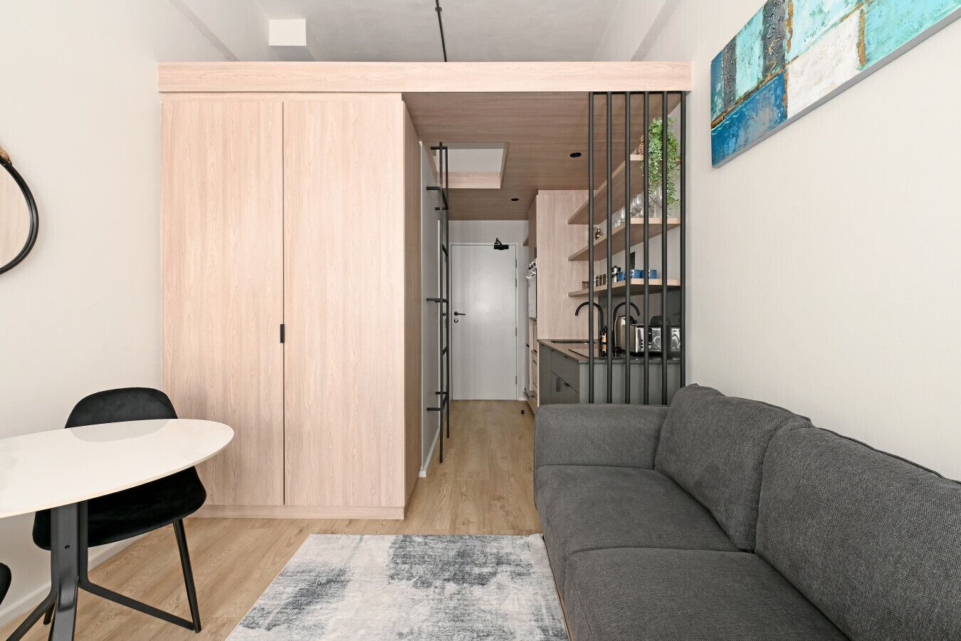 A compact living space with a kitchenette, a grey sofa, a dining area with a table and chairs, and a light wooden wardrobe.