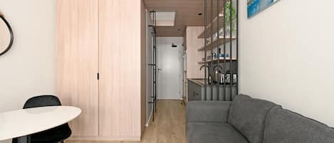 A compact living space with a kitchenette, a grey sofa, a dining area with a table and chairs, and a light wooden wardrobe.