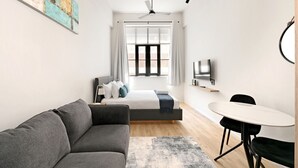 Una habitación de hotel moderna con un sofá gris, una cama con ropa de cama blanca y azul, una mesita redonda pequeña, un televisor en la pared y un ventanal con cortinas.