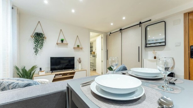 Un salon moderne doté d’un téléviseur à écran plat, d’un coin repas avec des assiettes et des verres, et d’une cuisine visible au fond.