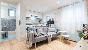 Un salon moderne avec un canapé gris, deux petites tables rondes blanches et une cuisine en arrière-plan.