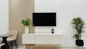 Een moderne woonkamer met een flatscreen televisie aan de muur, een witte console tafel, een ronde eettafel, een beige fauteuil en twee potplanten.