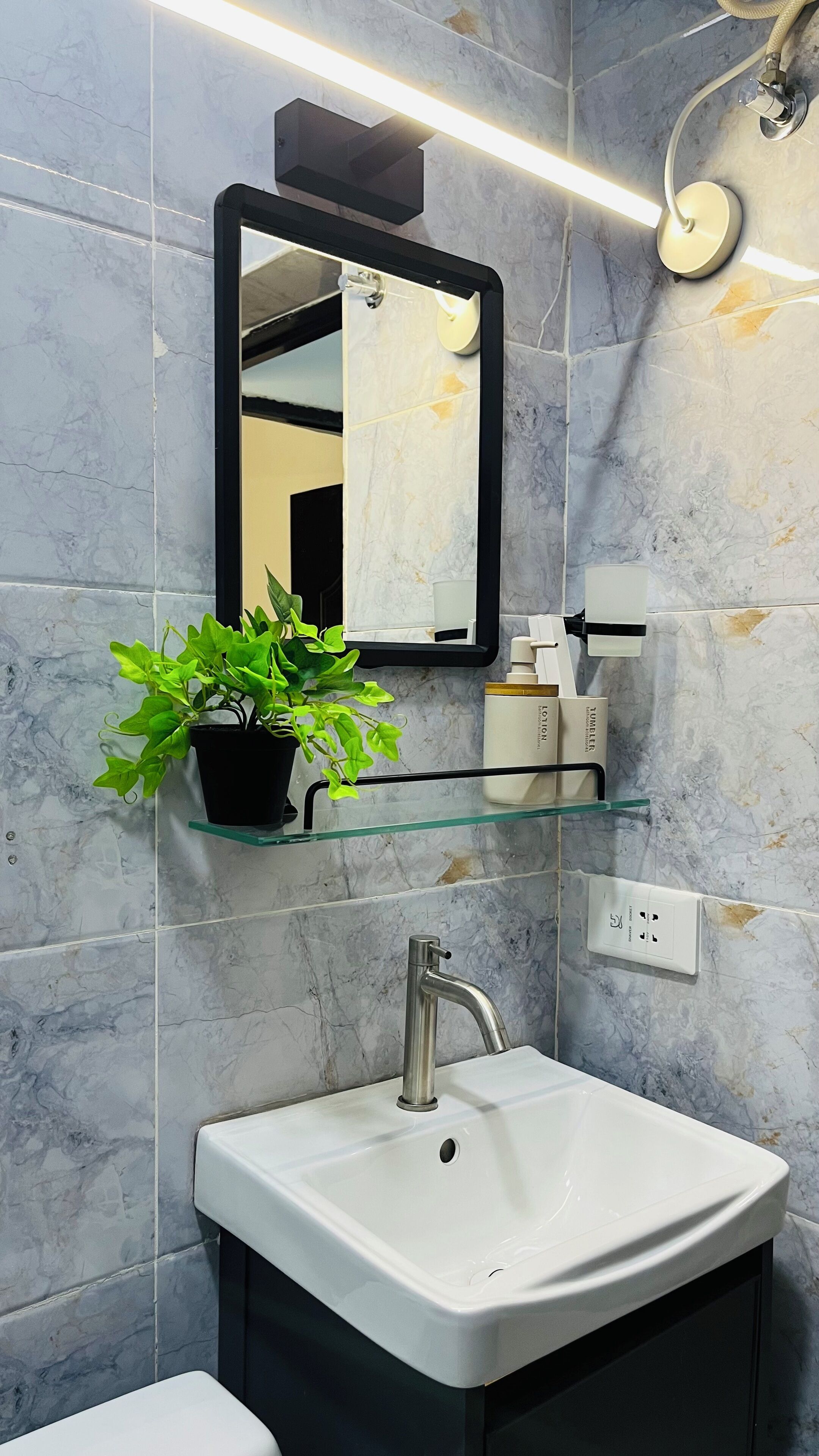 A bathroom with a white sink, a mirror, a potted plant, and toiletries on a shelf.