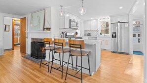A modern kitchen with white cabinets, a central island with bar stools, a fireplace, and a large window. - Coastal comfort near beaches and walking distance to Harwich Port! (Harwich)