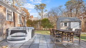 A patio with a stone floor, a hot tub, and a wooden table with chairs. - Coastal comfort near beaches and walking distance to Harwich Port! (Harwich)