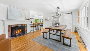 A modern living room with a fireplace, wooden flooring, a dining table with benches, and a large window. - Coastal comfort near beaches and walking distance to Harwich Port! (Harwich)