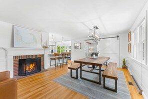 A modern living room with a fireplace, wooden flooring, a dining table with benches, and a large window. - Coastal comfort near beaches and walking distance to Harwich Port! (Harwich)