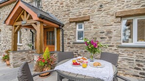 A stone building with a wooden porch, a table with breakfast items, and potted plants. - 3 Bed in Ponthirwaun (oc-t33294) (Ponthirwaun)
