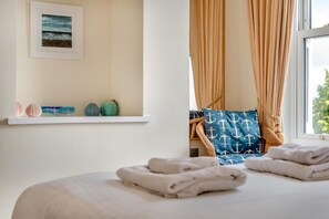 A neatly made bed with folded towels, a shelf with decorative items, a framed picture of the sea, and a window with curtains. - 3 Bed in Fowey (oc-o33078) (Fowey)