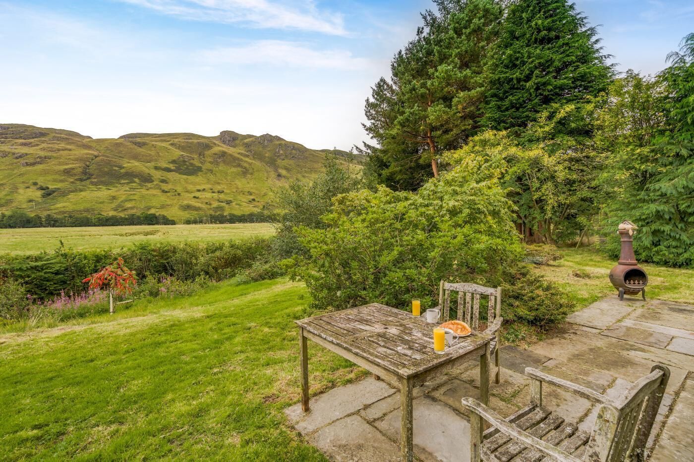 Un espace extérieur aménagé dans un style rustique, avec une table et un banc en bois, entouré d’une végétation luxuriante et offrant une vue sur des collines ondulantes.