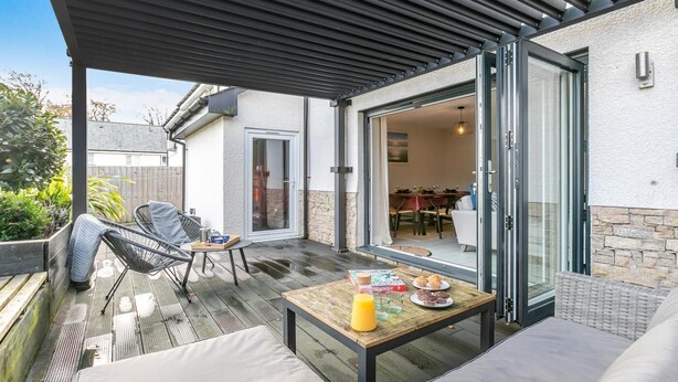 An outdoor patio with a wooden table, a sofa, and a chair. There is a glass door leading to an indoor dining area. - 3 Bed in Fowey (oc-s33234) (Fowey)